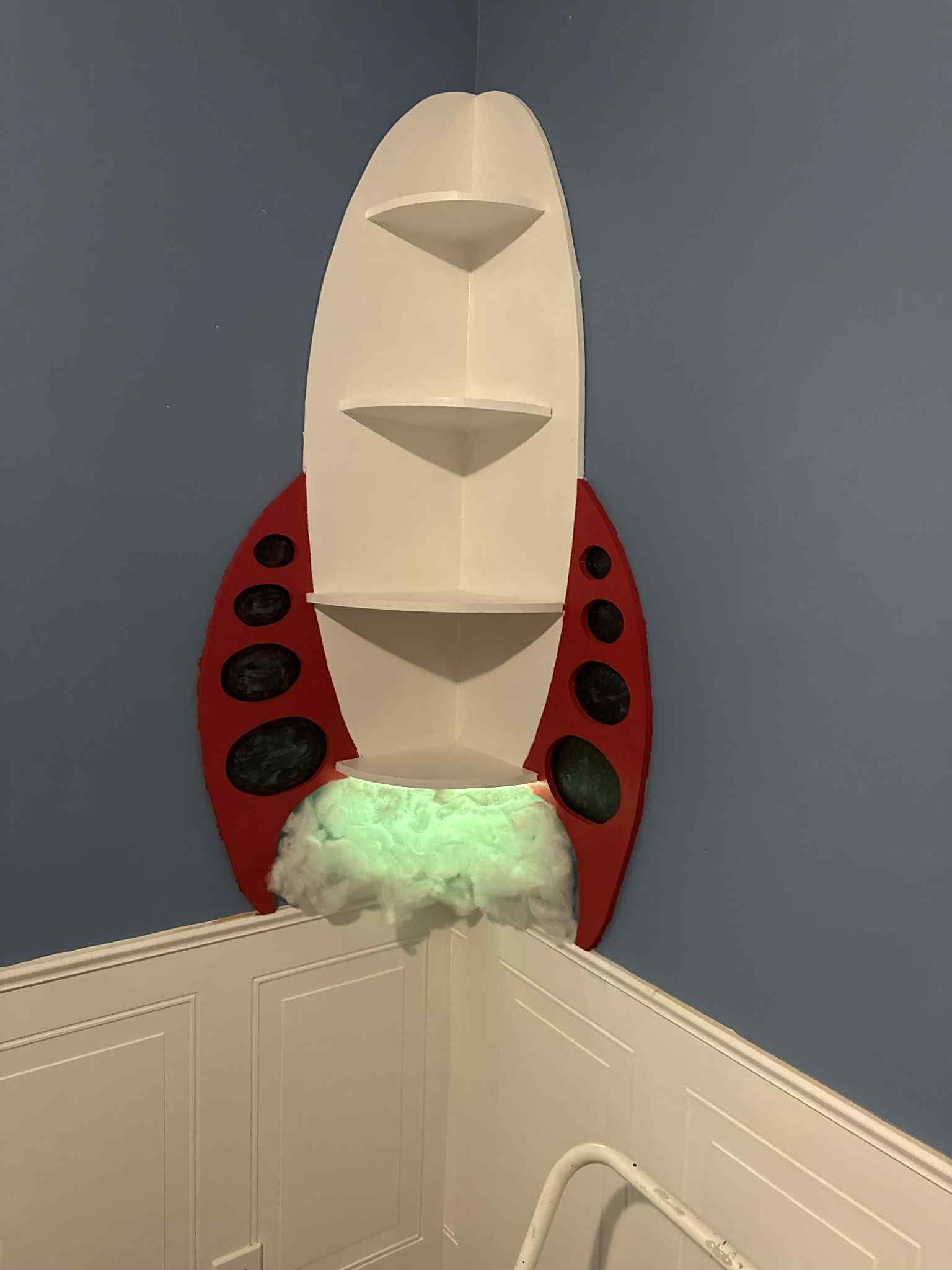 A rocket-shaped shelf on a blue wall. Red body with white top and three shelves. Green glowing