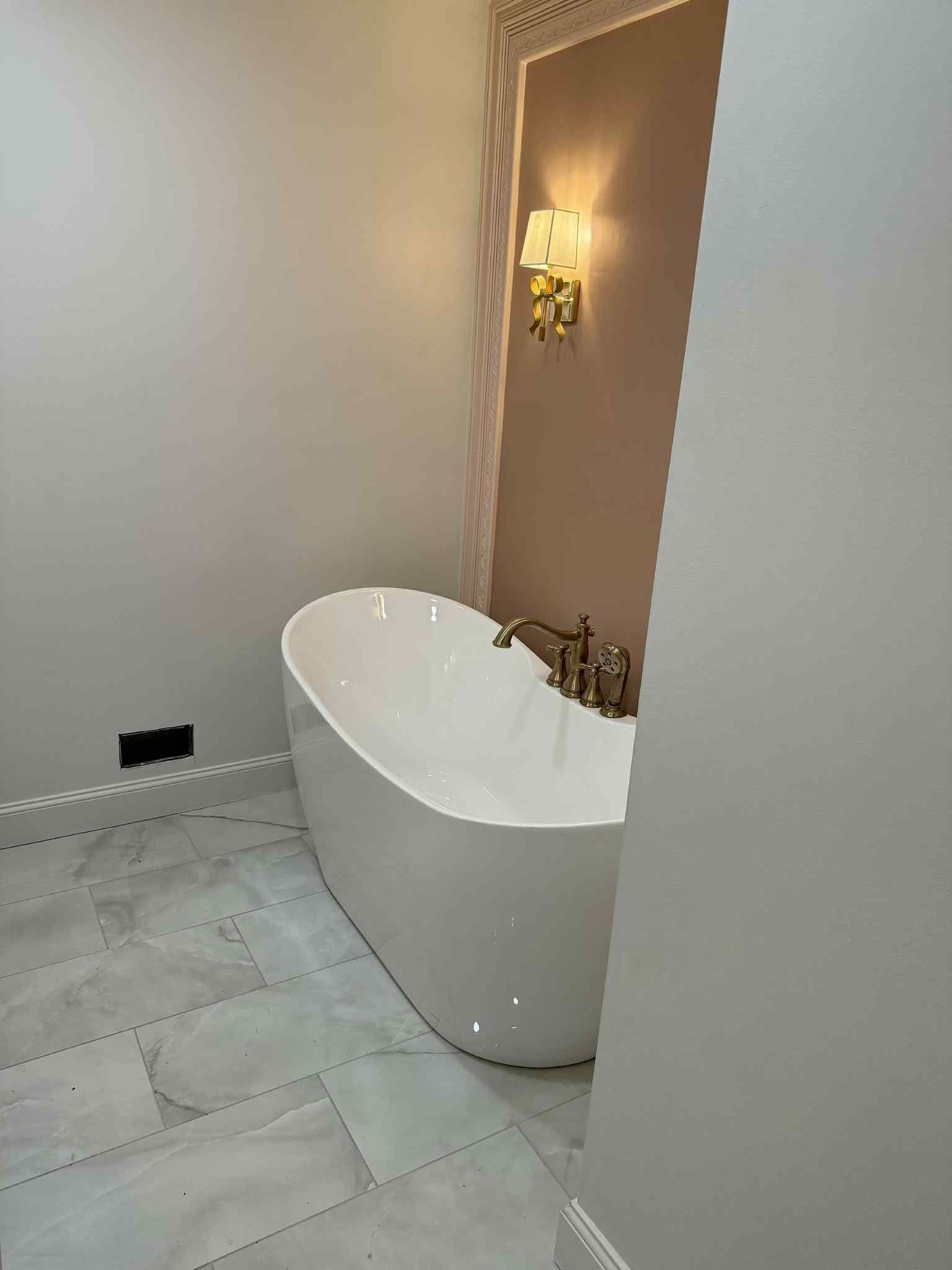 Freestanding white bathtub in a bathroom with pale walls and marble floor. Golden sconce.