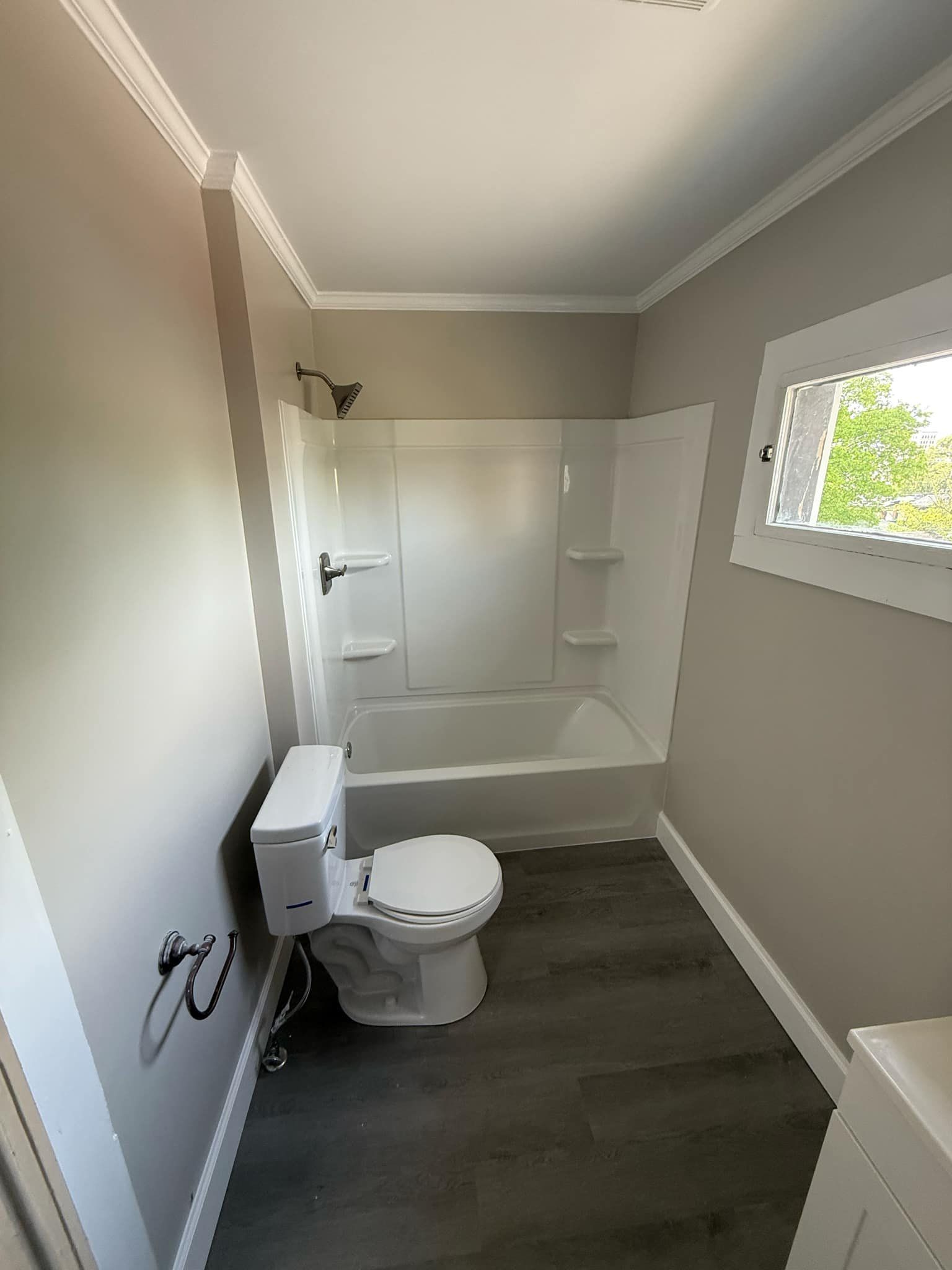 Bathroom with white tub/shower, toilet, and window, gray walls and flooring.