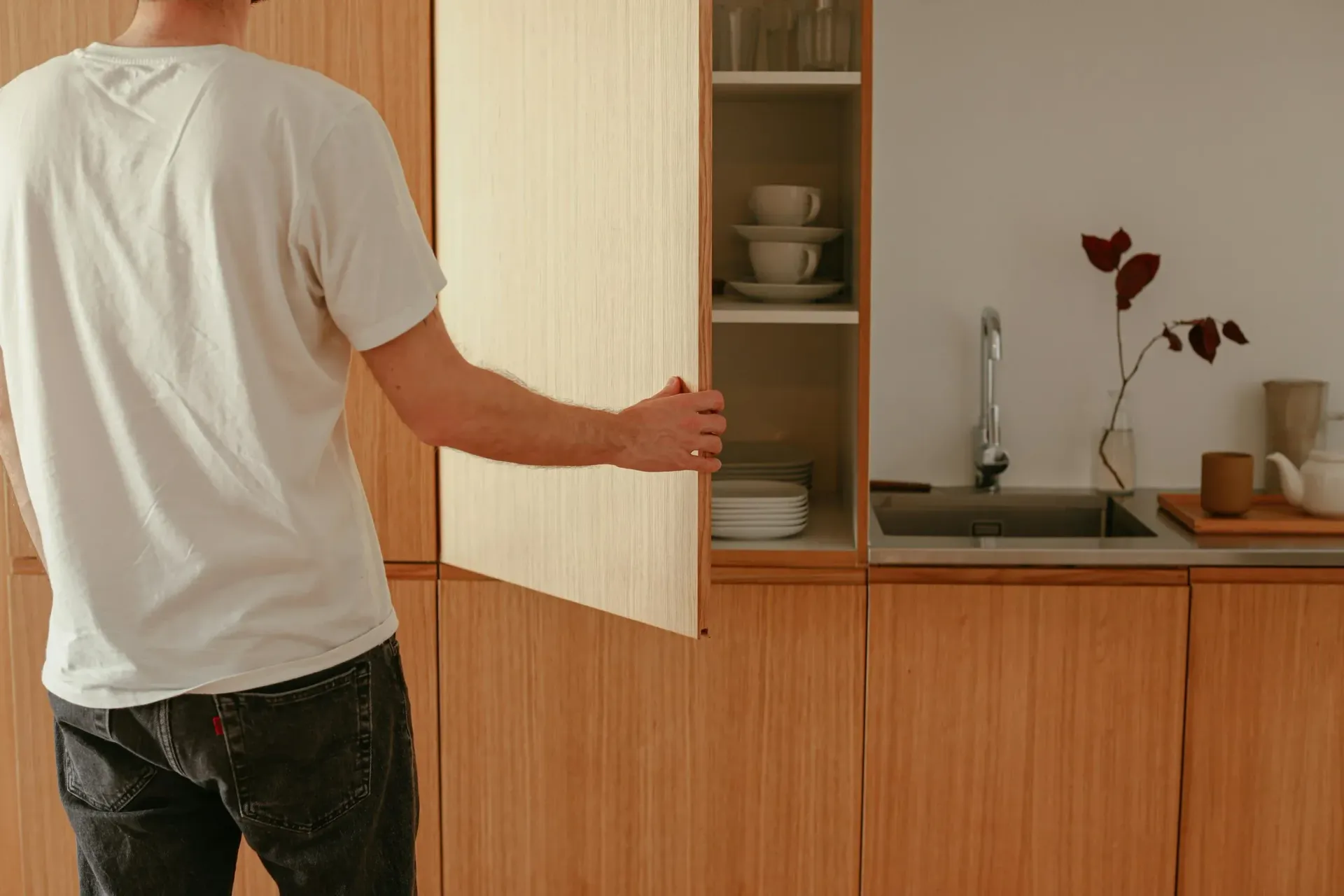 Person opening light wood kitchen cabinet, revealing dishes and sink.