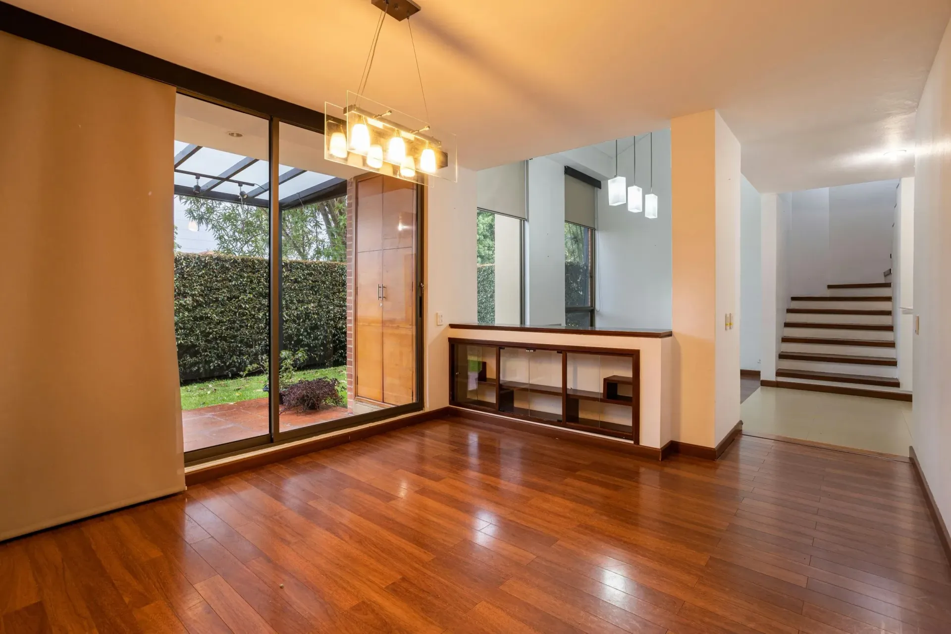 Empty dining room with wood flooring, large windows overlooking a green garden, and a staircase.