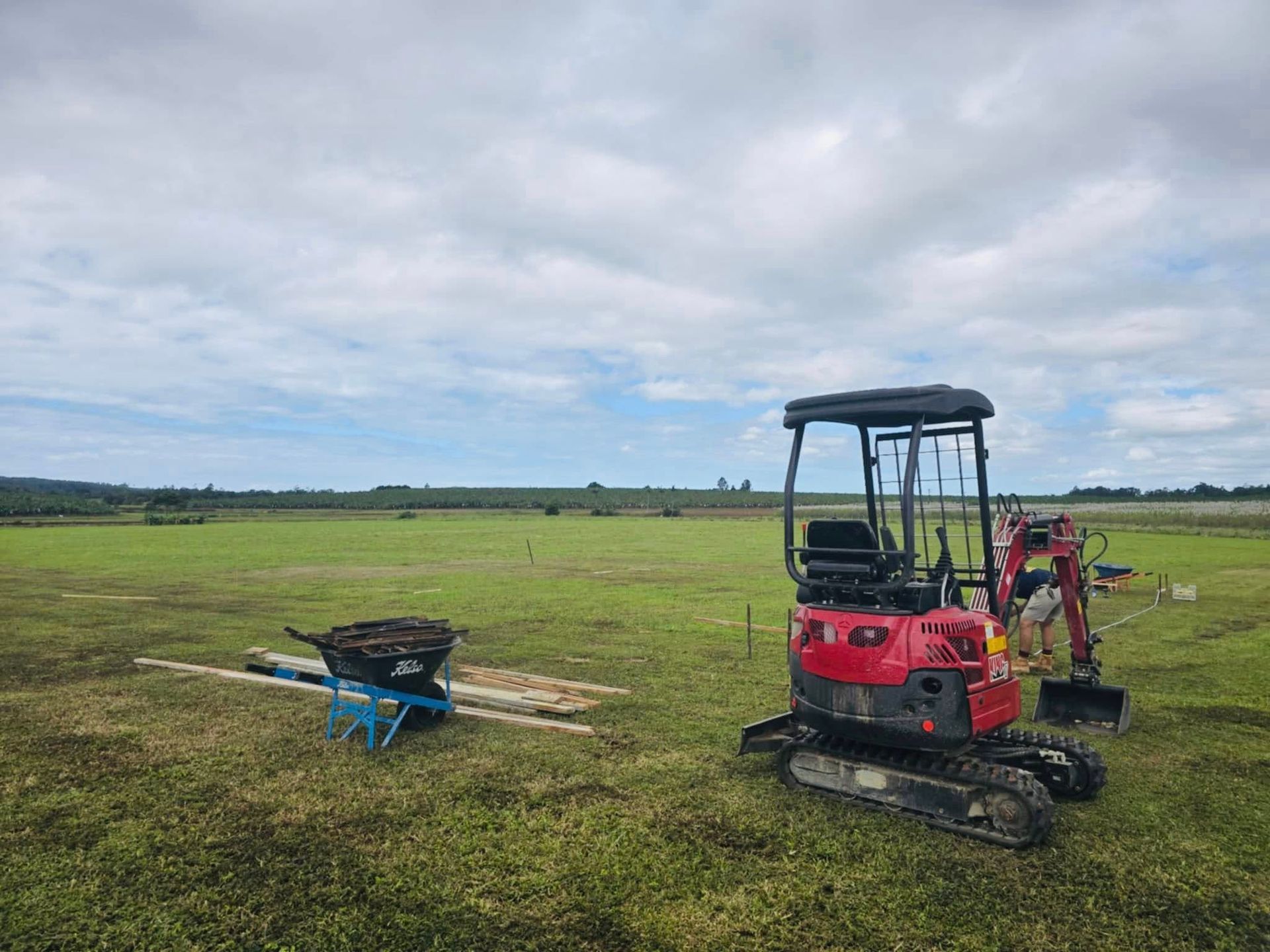 Red Excavator and Wheelbarrow on a Green Field Under a Cloudy Sky — Sun Co Concrete And Construction in Dimbulah, QLD