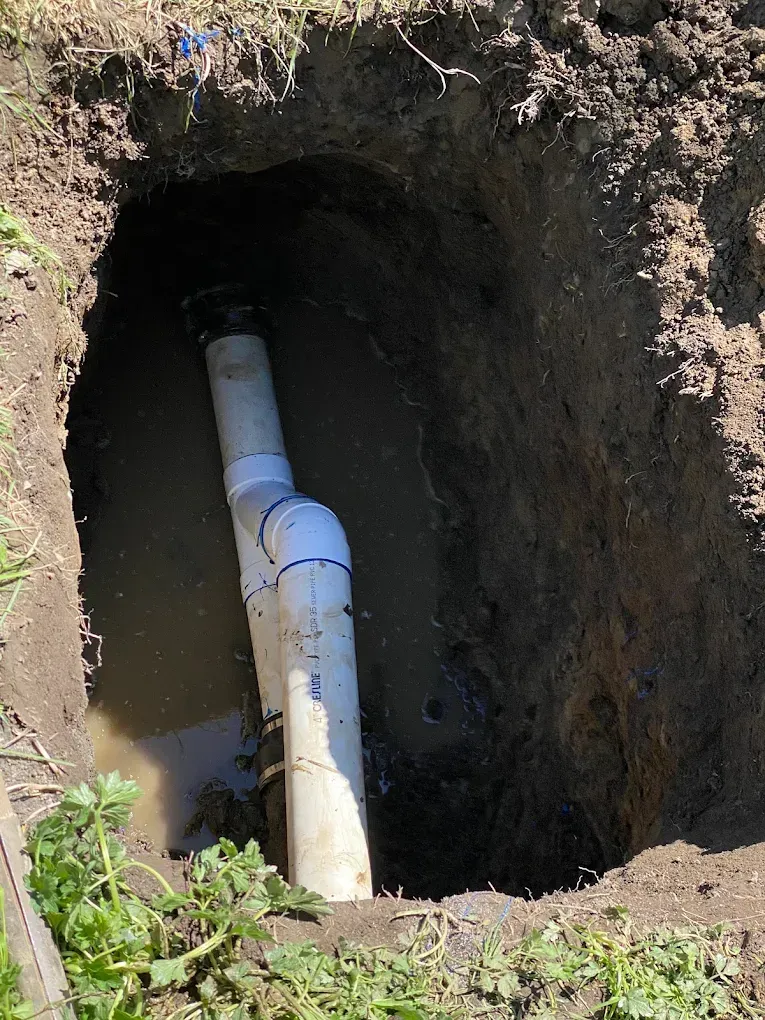 A hole in the ground reveals a white PVC pipe with water pooling inside, set in a grassy area.