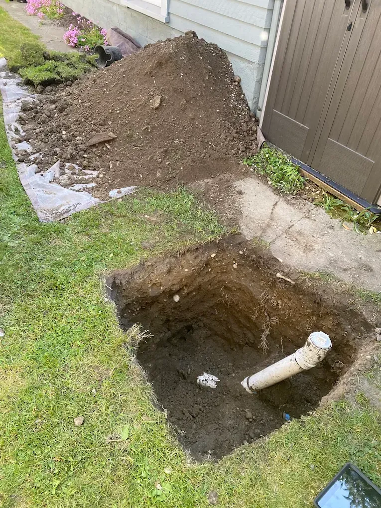 An open square hole in a grassy yard with a pipe inside, dirt pile nearby, next to a building.