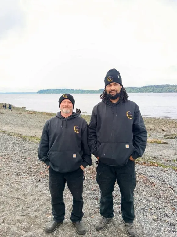 Two men in black hoodies and beanies stand on a rocky shoreline overlooking a body of water.