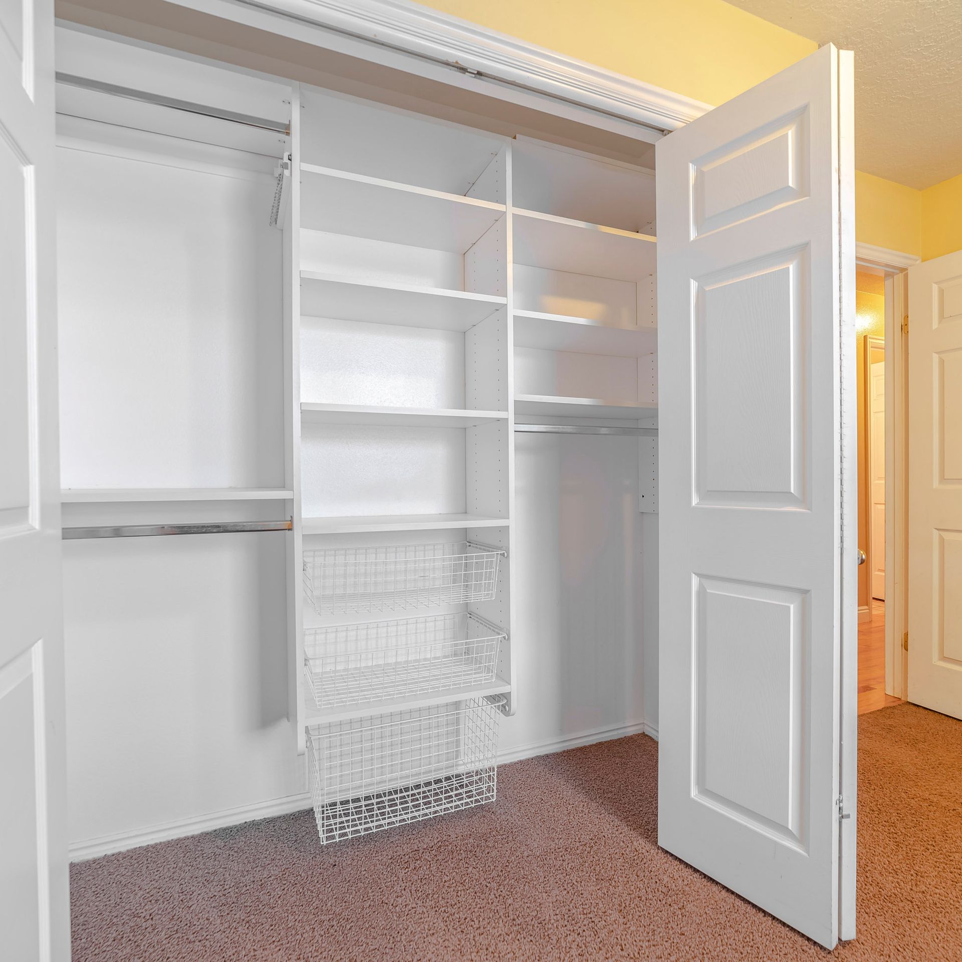 A walk-in closet with white shelving, a hanging rod, wire baskets, and white bi-fold doors over brown carpet.