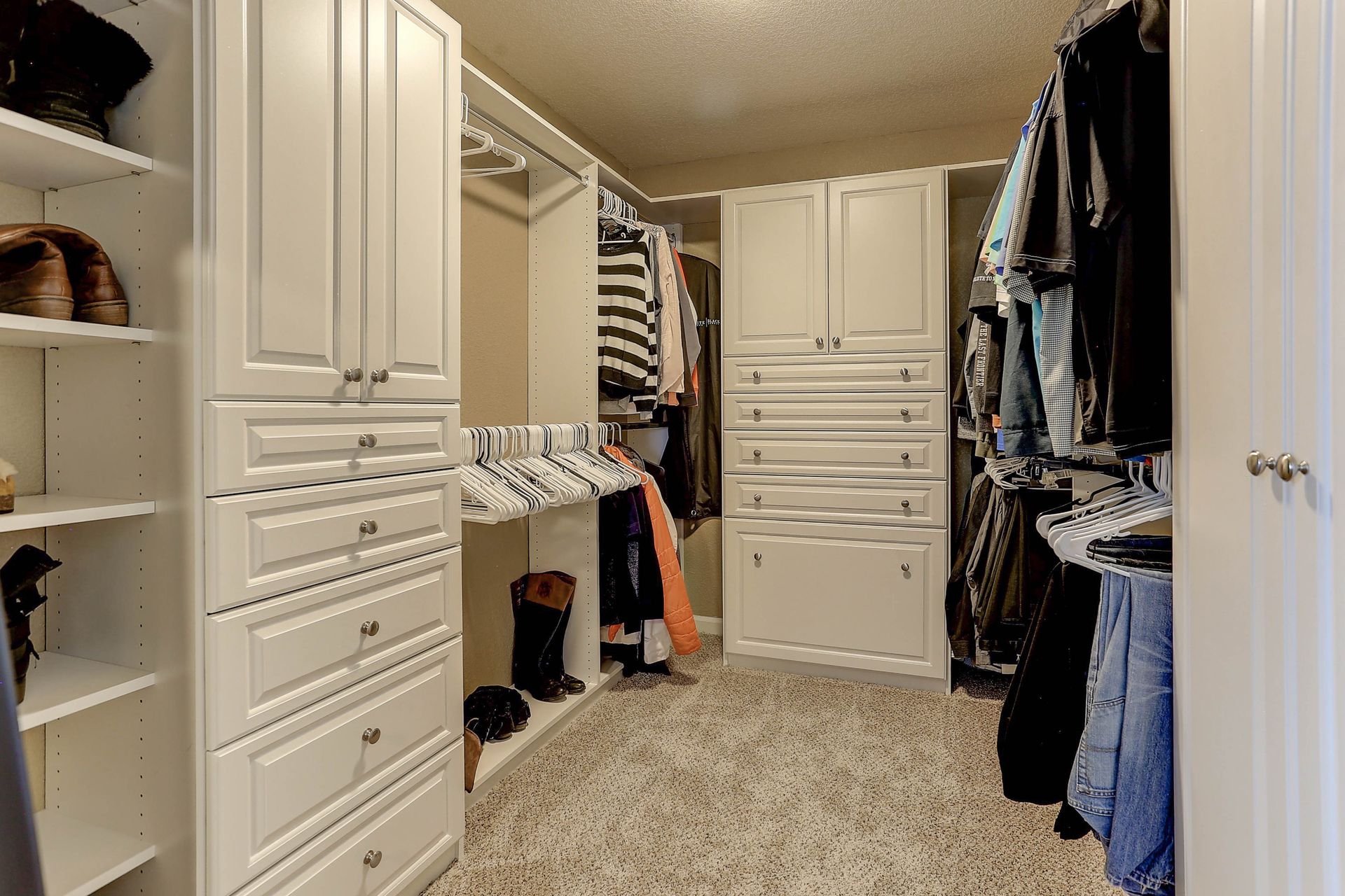 A walk-in closet featuring white wooden cabinetry, organized hanging clothing, and a beige carpeted floor.