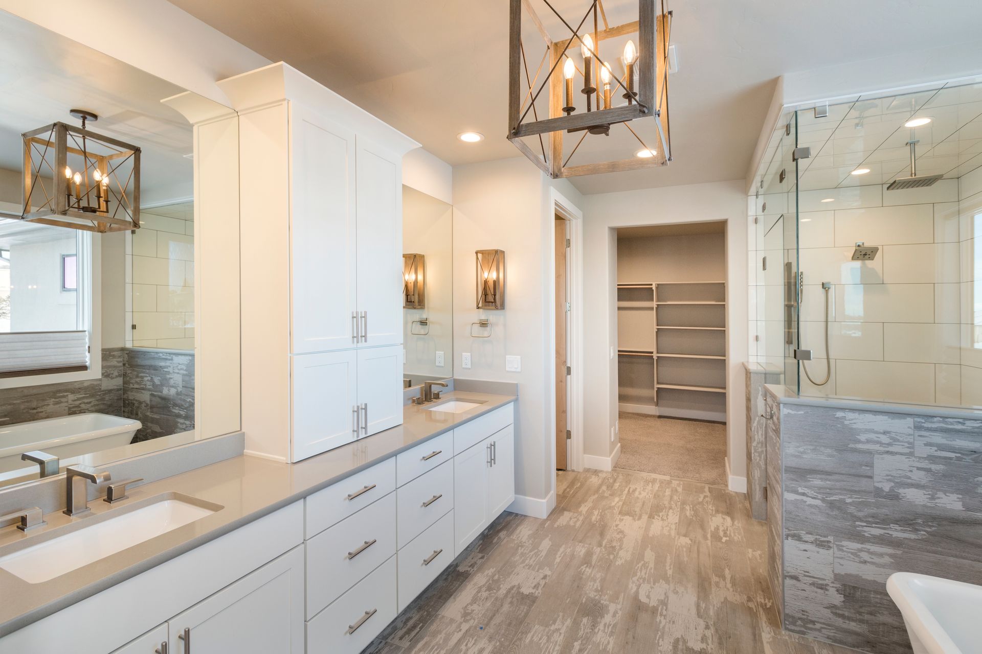 A modern bathroom with a double vanity, gray cabinets, dual mirrors, and a white soaking tub next to a window.
