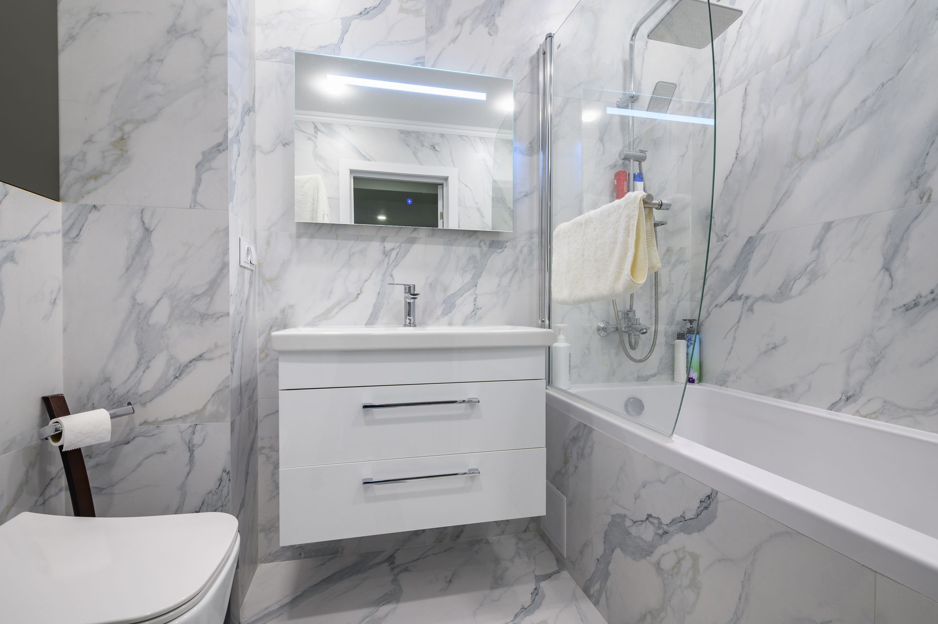 A modern bathroom with white marble-patterned tiles, a floating vanity, a toilet, and a glass-enclosed bathtub shower.