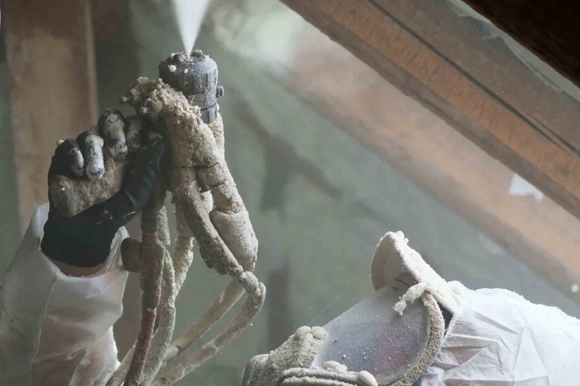 Person spraying insulation into an attic, wearing protective gear.