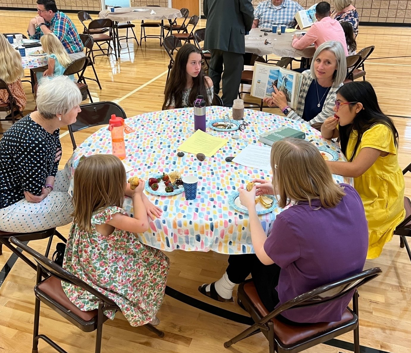 People seated around a decorated table, eating and reading, in a community setting.