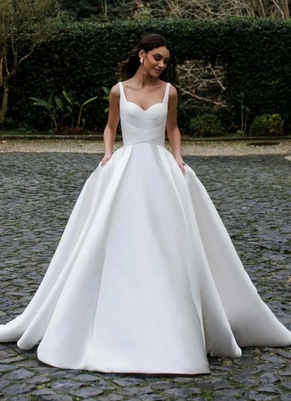Woman in a white ball gown standing on a cobblestone path outdoors, smiling softly.