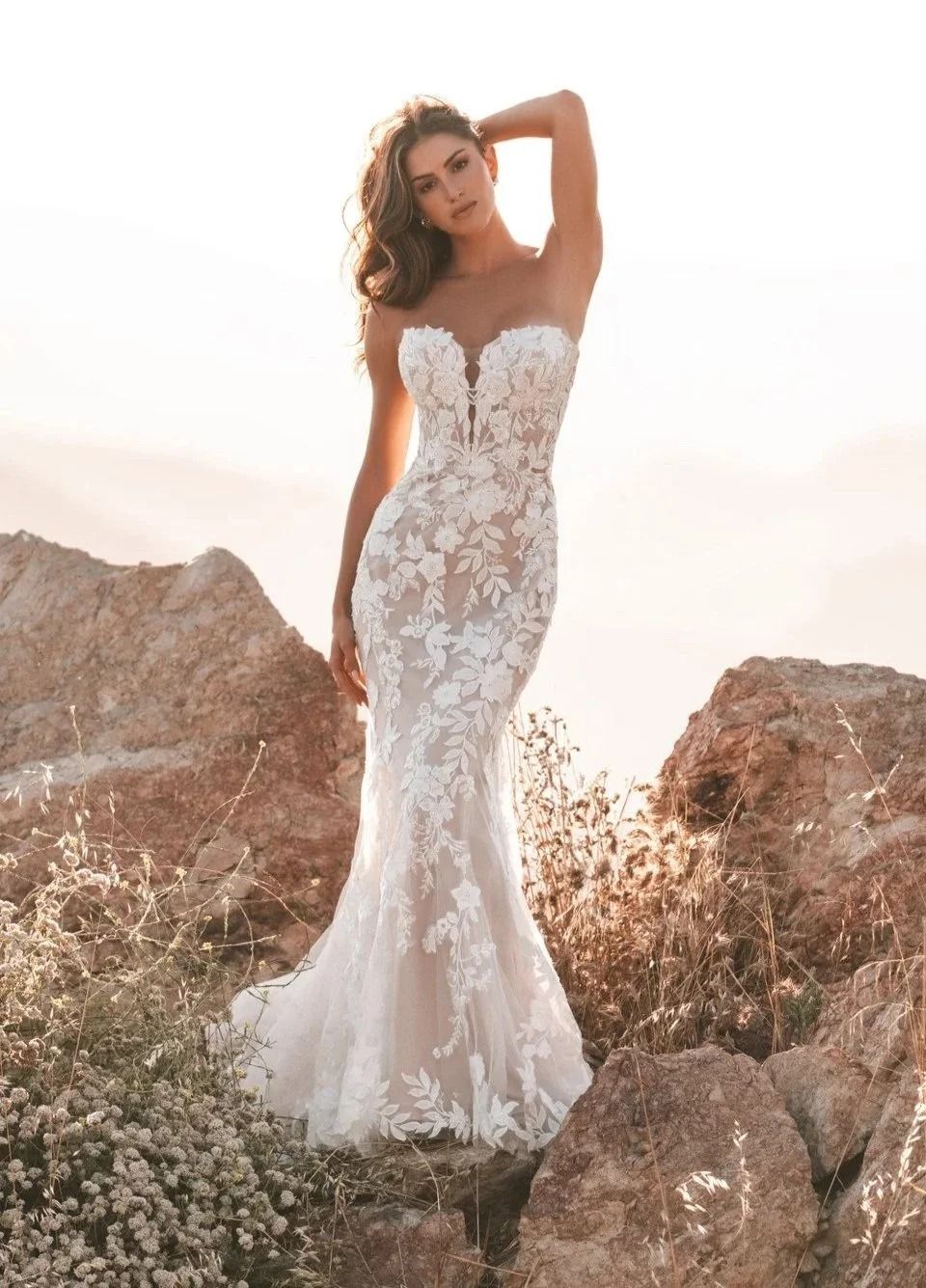 Bride in a white lace mermaid gown standing among rocks in a sunlit field