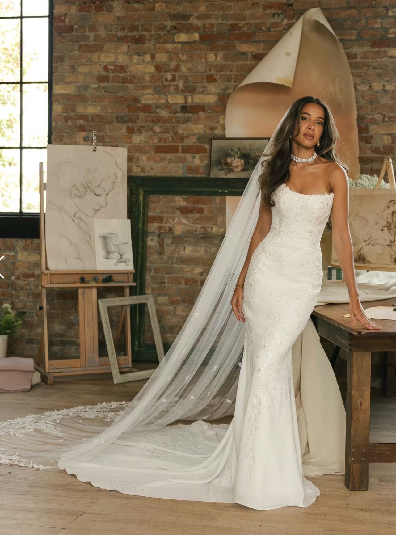 Bride in white mermaid gown with long veil standing in a rustic studio with brick walls and easel paintings