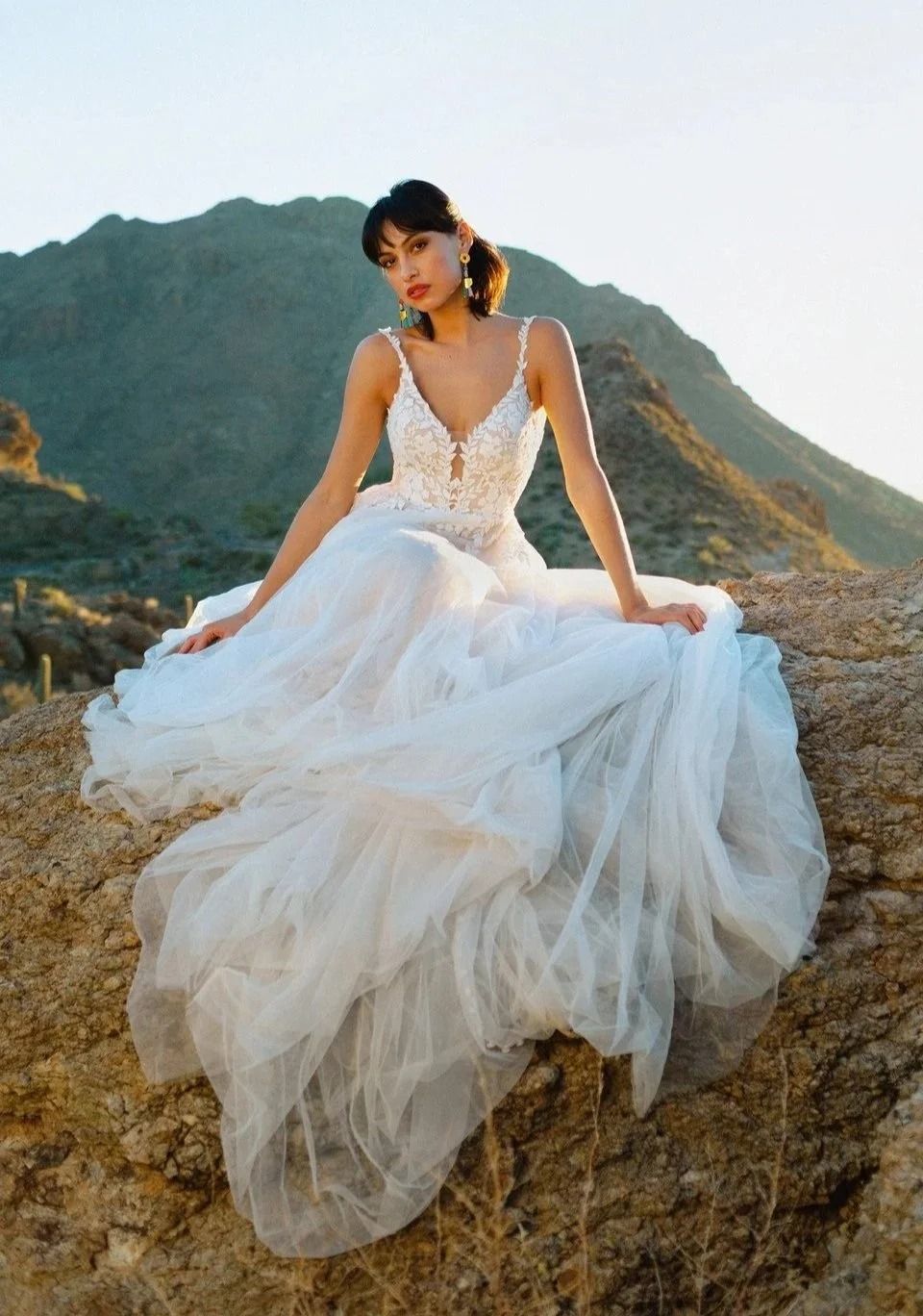Woman in a white tulle gown seated on a rocky cliff with mountains in the background.