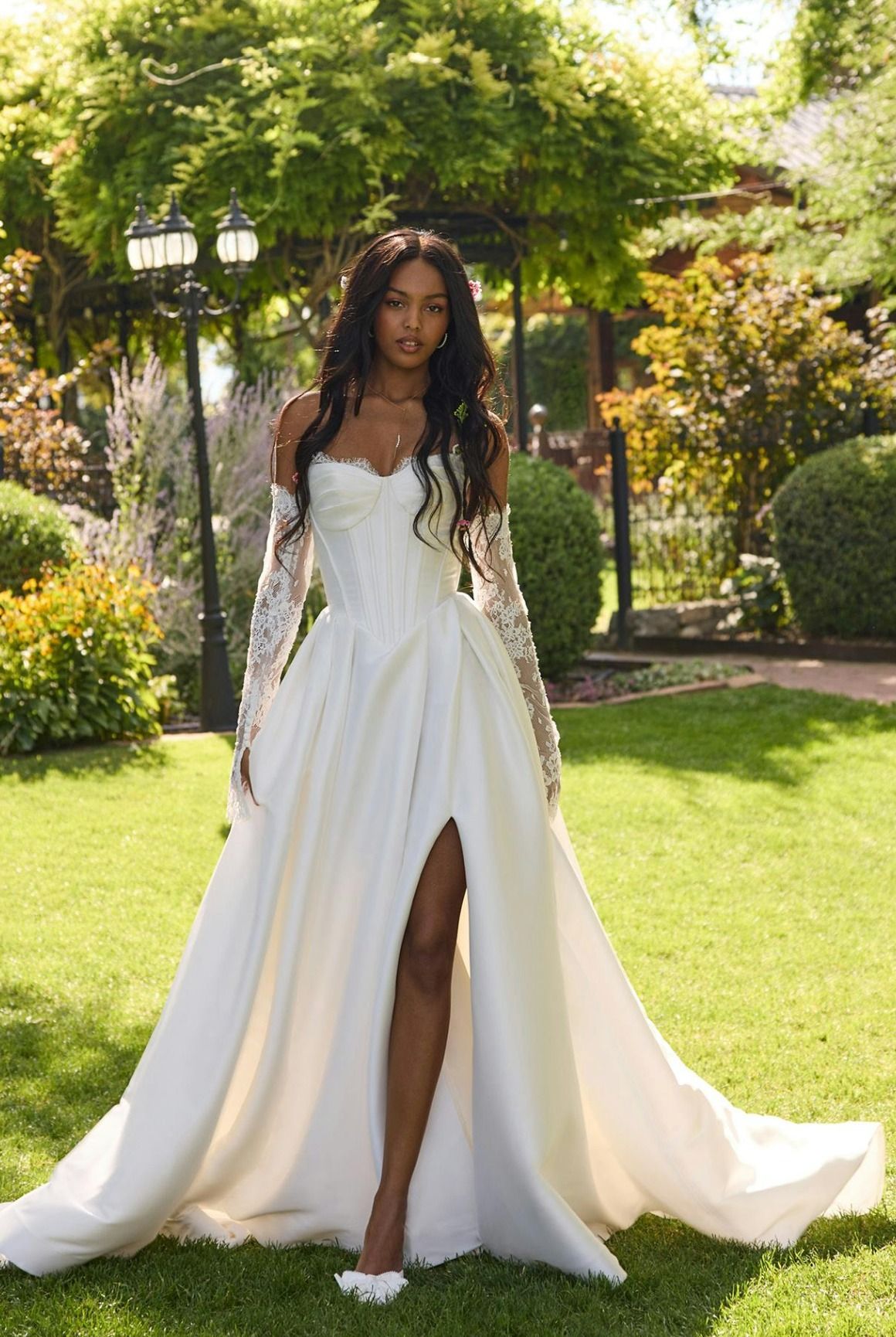 Woman in a white gown standing on a sunlit garden lawn with trees and flowers.