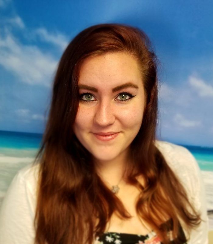 A woman with long red hair is smiling in front of a beach background