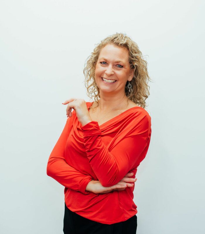 A woman in a red shirt is standing with her arms crossed and smiling.
