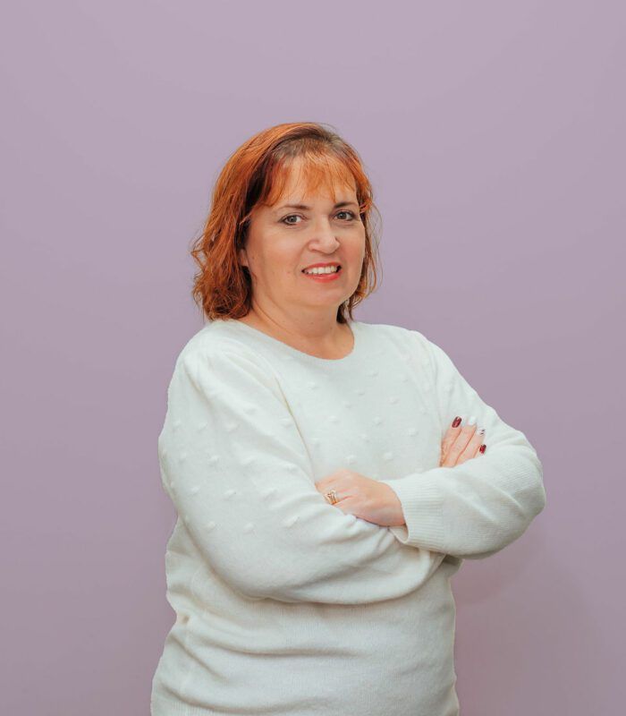 A woman with red hair is wearing a white sweater and standing with her arms crossed.
