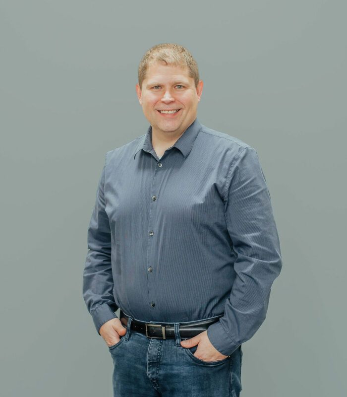 A man in a blue shirt and jeans is standing with his hands in his pockets.