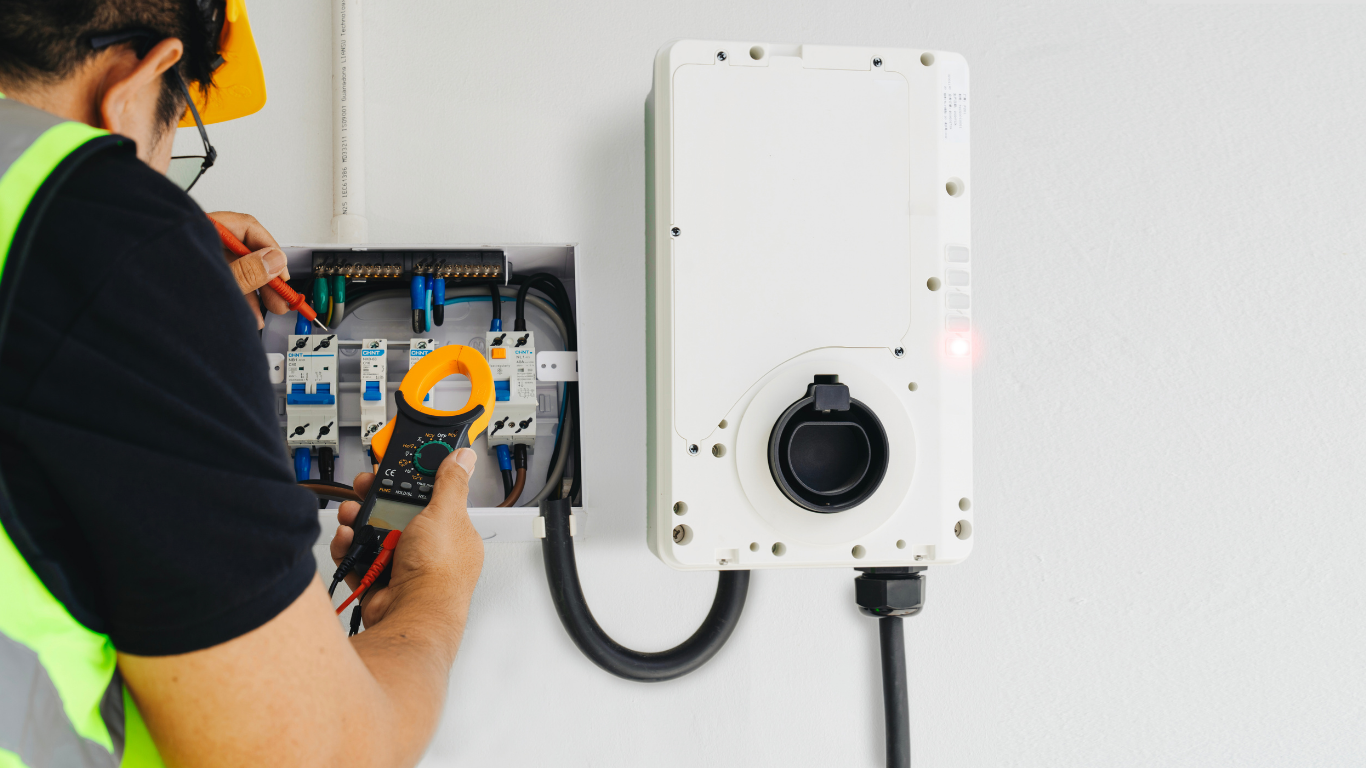 Electrician using a multimeter to inspect wiring inside an electrical panel near a charging station.