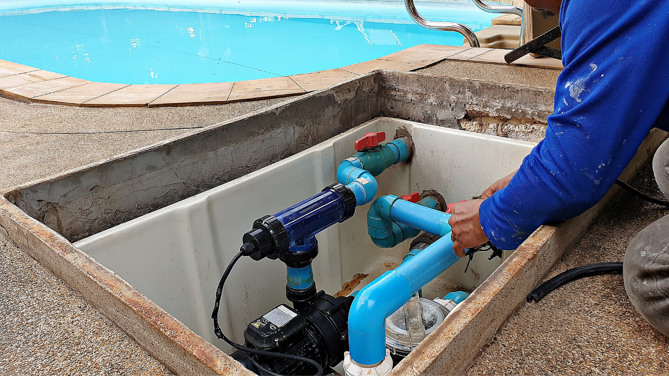 Person working on pool equipment in a concrete enclosure. Blue pipes and pump visible. Pool in background.