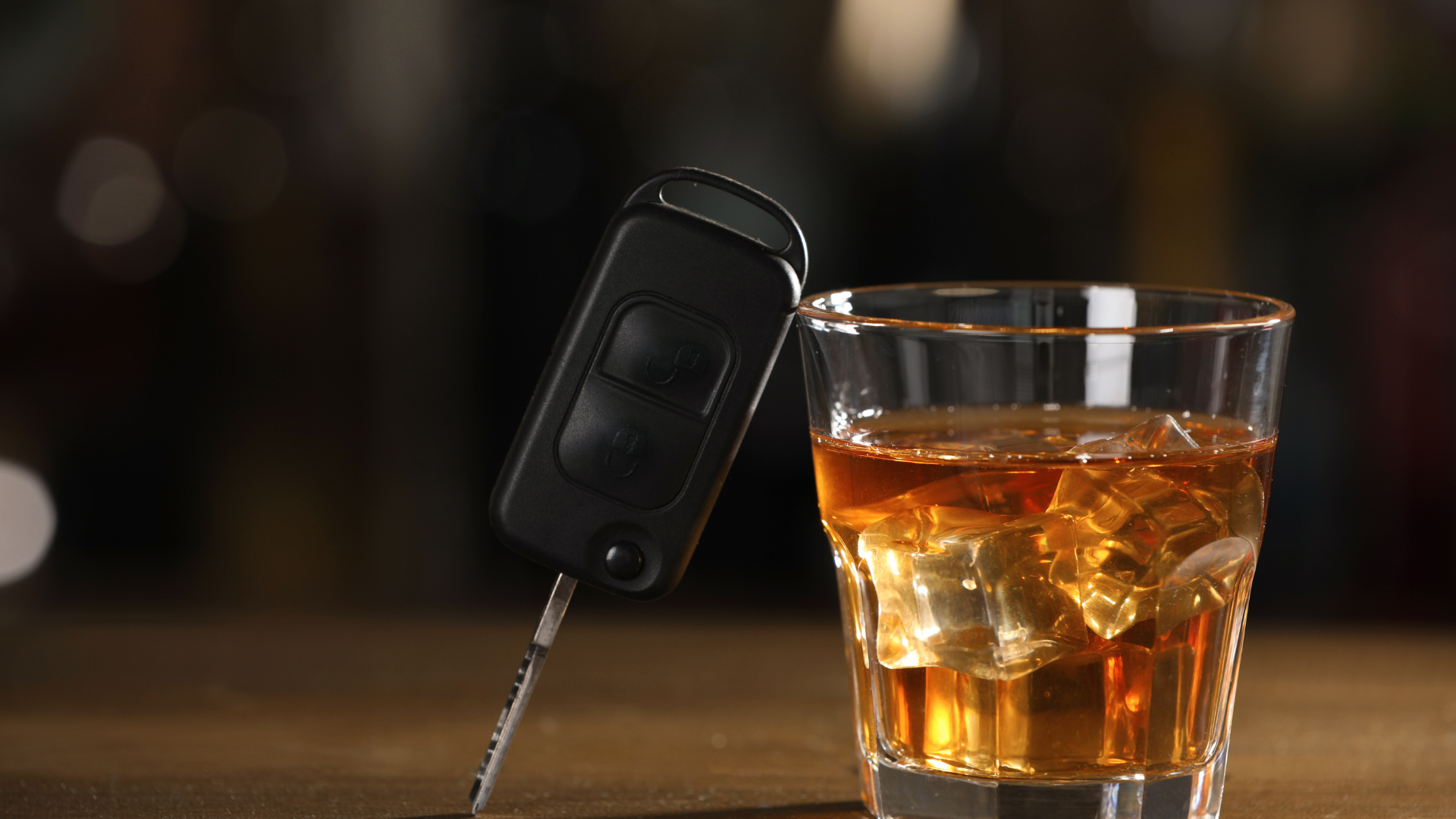 A car key fob rests against a glass of amber-colored liquid with ice, set on a wooden surface against a dark background.