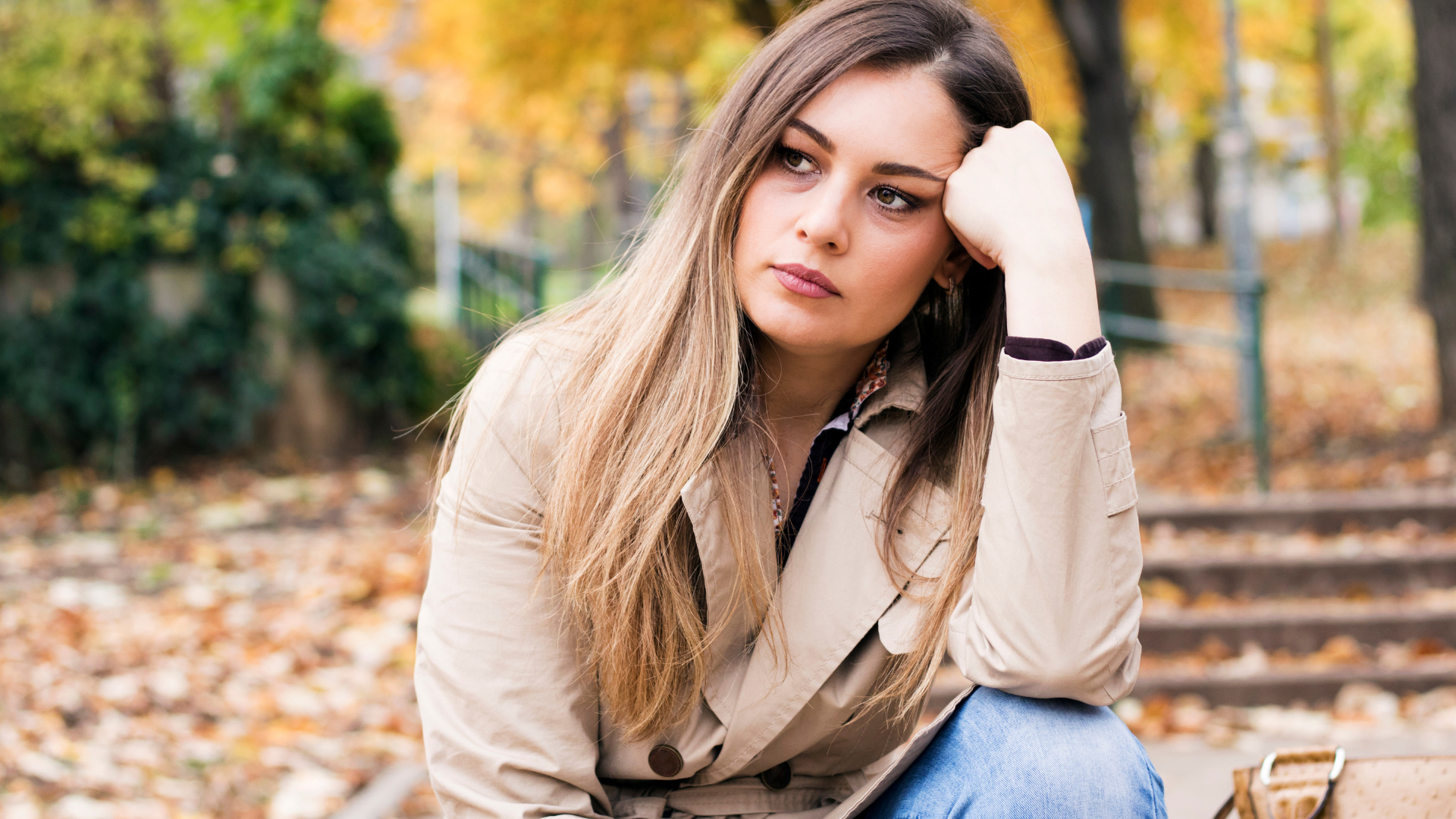 A person with long blonde hair rests their head on a hand while sitting outdoors in an autumn park.