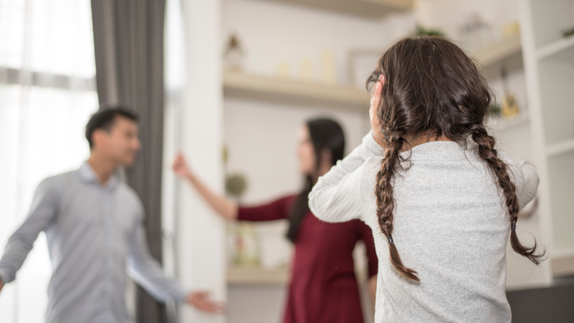 A child with braids covers their ears while a man and woman argue in the background of a blurred room.