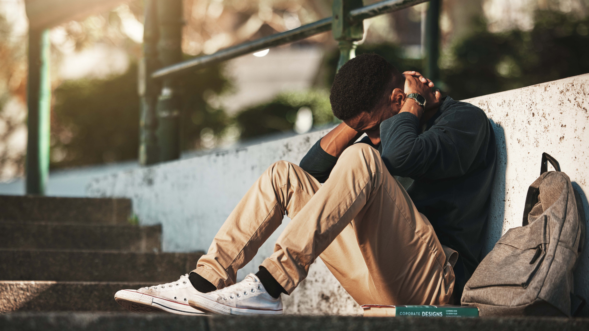 A person sits on outdoor stairs, leaning against a wall with their head in their hands and a backpack beside them.
