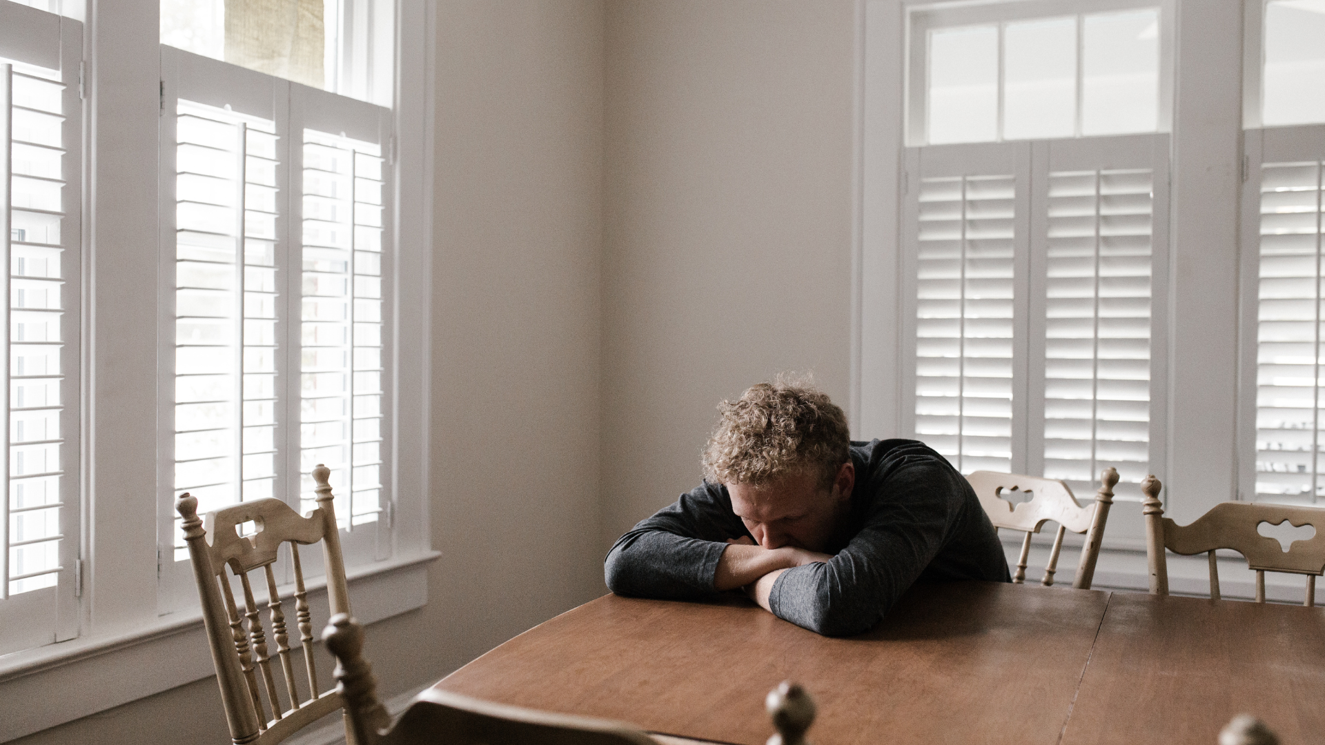 A person sits slumped over at a wooden dining table, resting their head on their crossed arms in a brightly lit room.