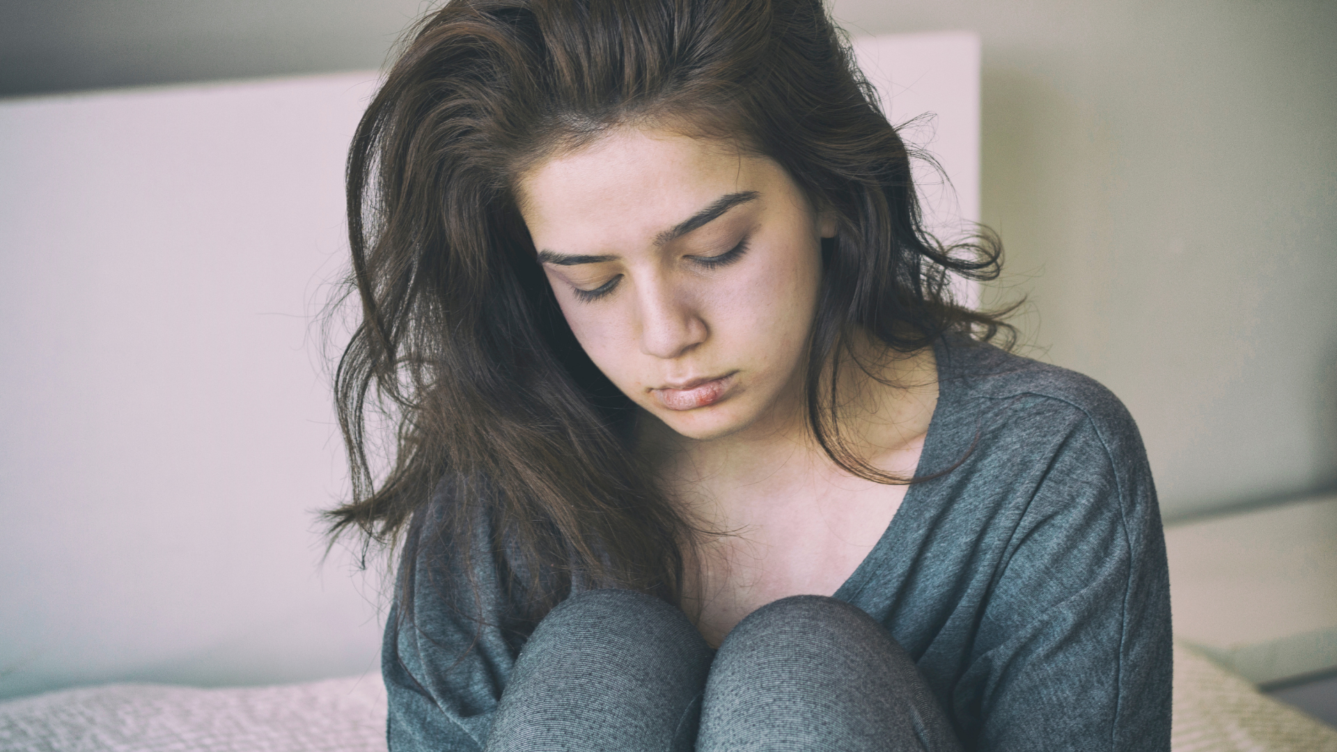 A person with long dark hair sits on a bed, looking downward with a neutral, contemplative expression.