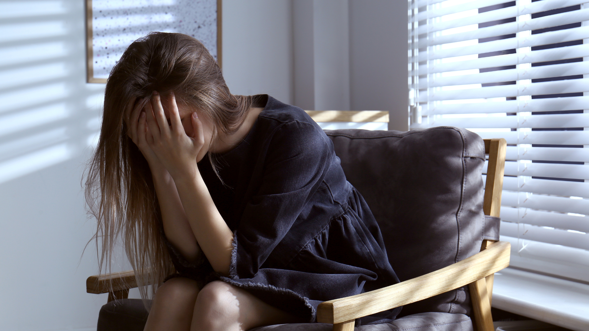 A person sits in a chair by a window with blinds, covering their face with their hands in a gesture of distress.
