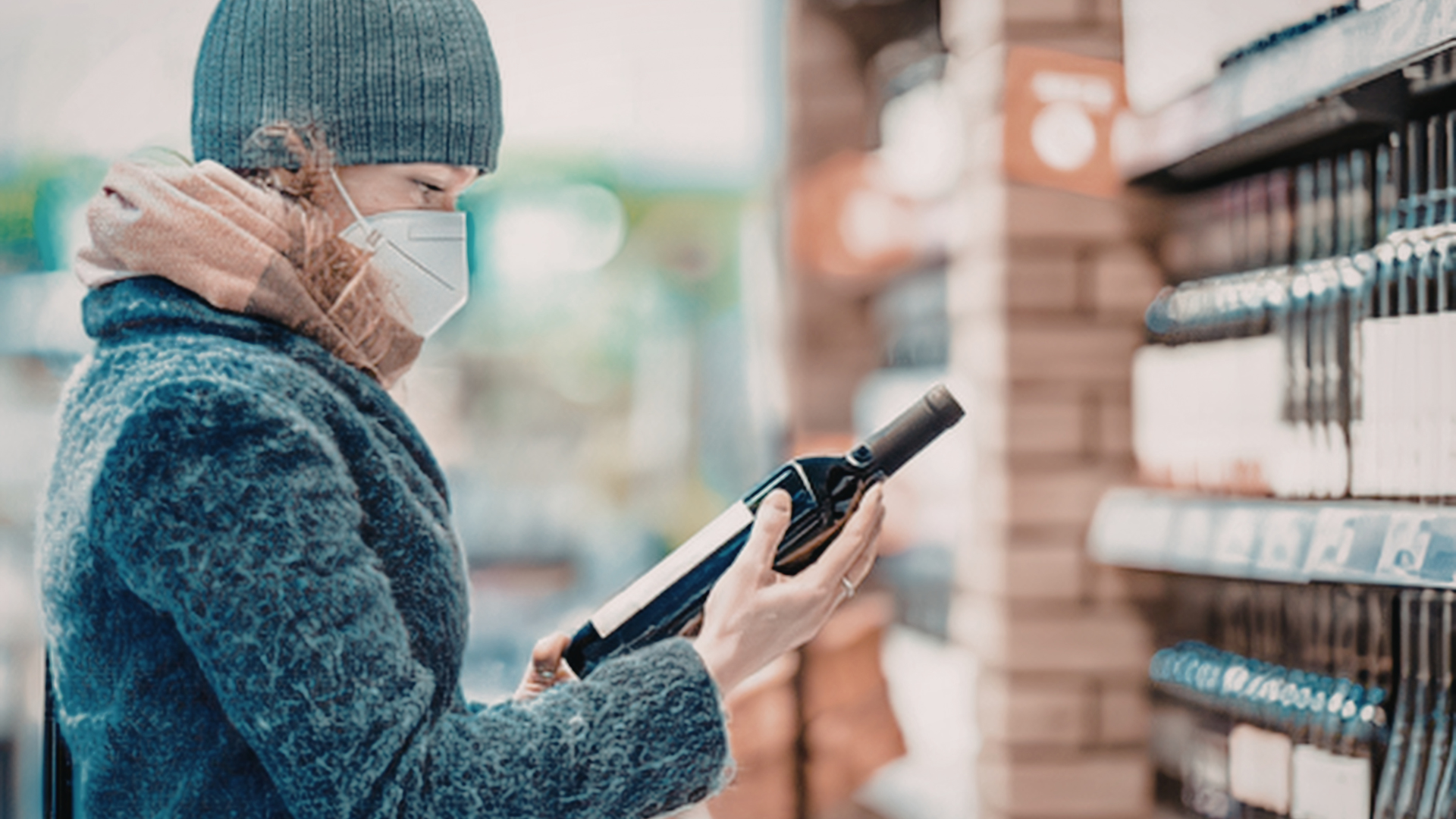 A person wearing a beanie, scarf, and face mask examines a wine bottle in a store aisle.