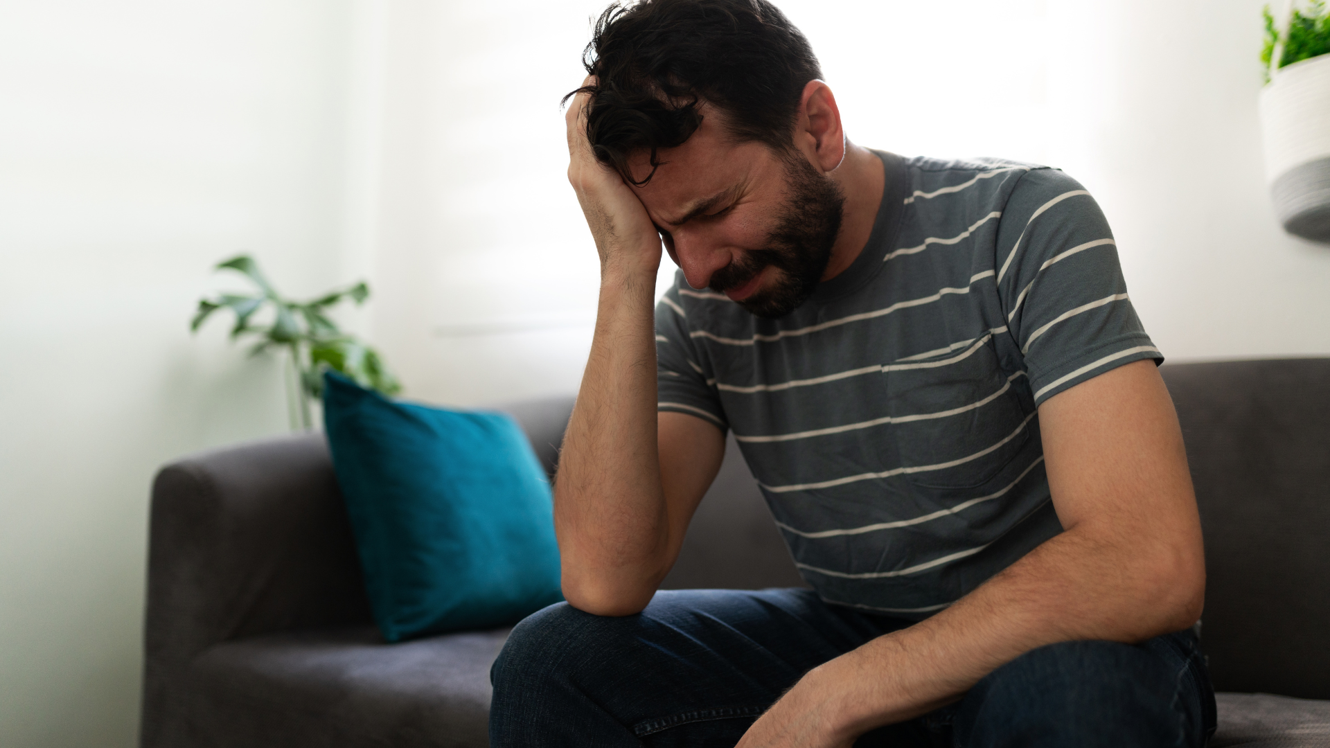 A person with a beard sitting on a grey couch, head in their hand, looking distressed.