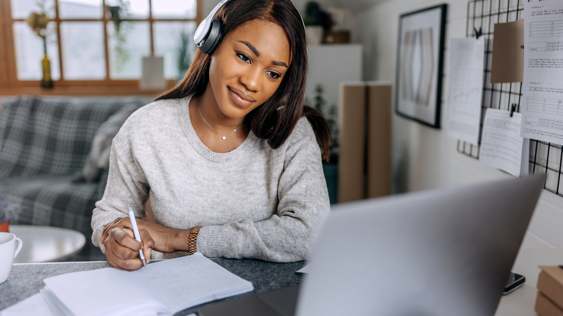 A person wearing headphones works at a desk, looking at a laptop while taking notes on paper in a bright home office.