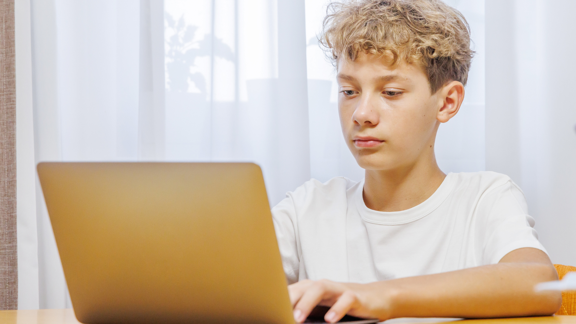 A person with light brown curly hair wearing a white t-shirt uses a laptop while sitting indoors.