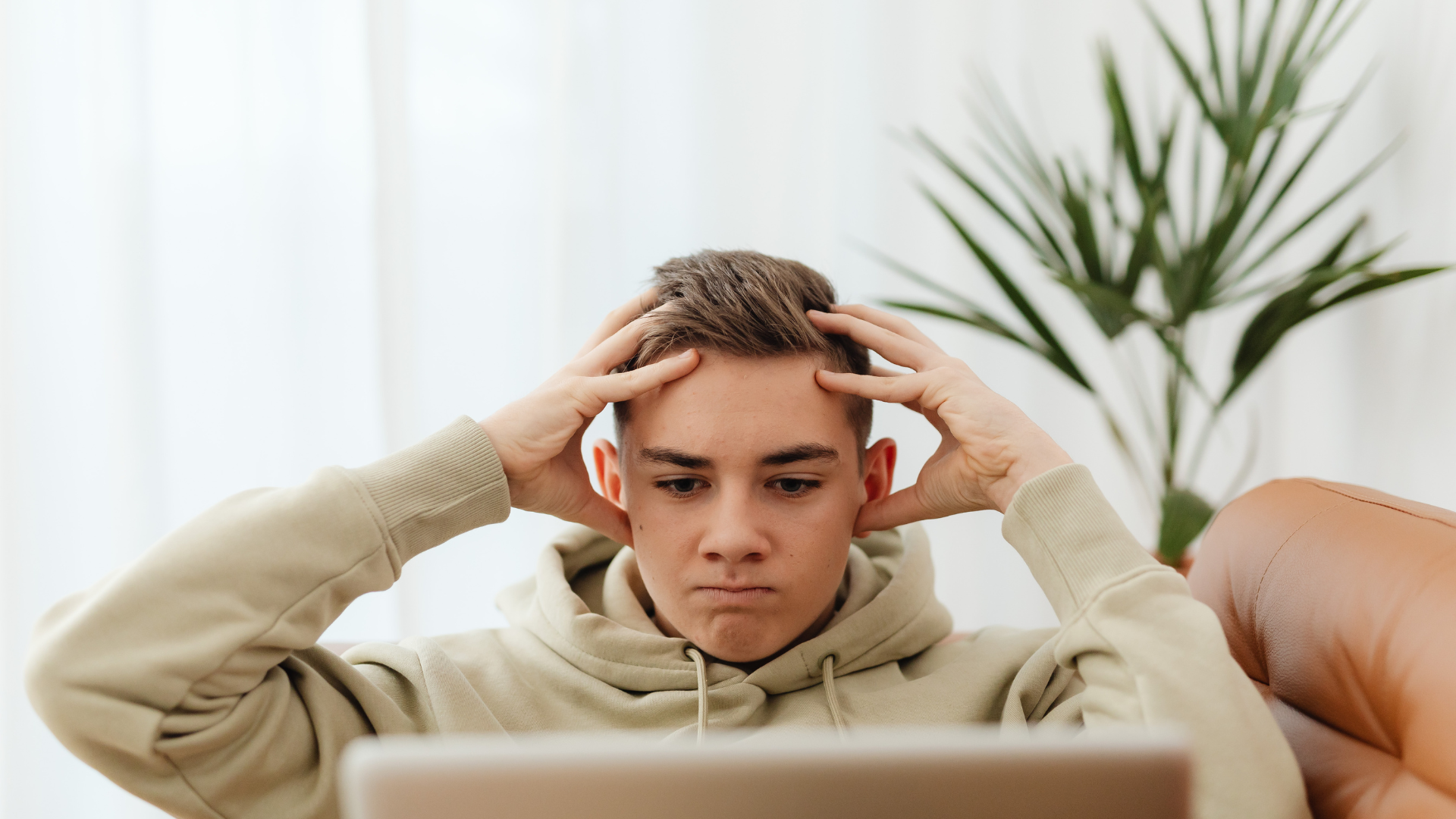 A person in a beige hoodie sits before a laptop with their hands on their head, looking frustrated or stressed.