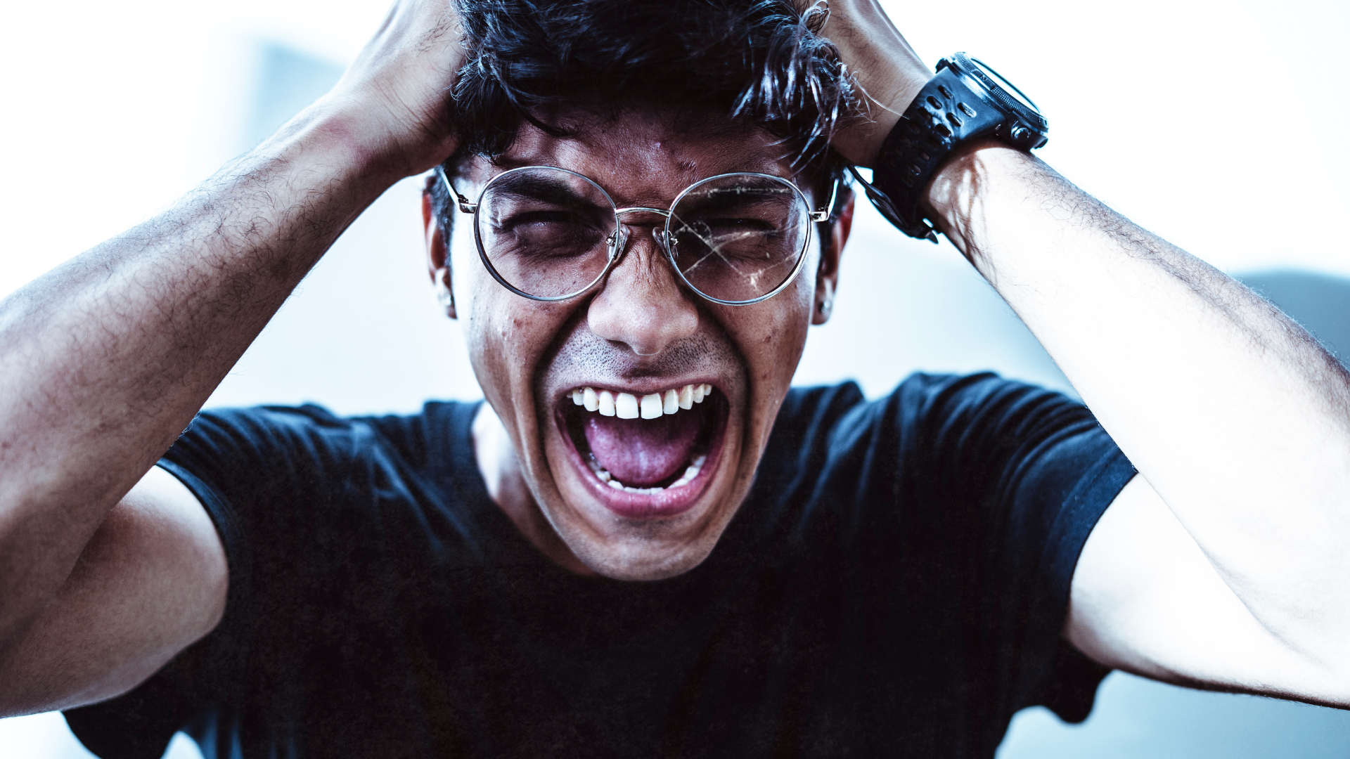 A person with messy hair and round glasses clutches their head while screaming with their mouth wide open.
