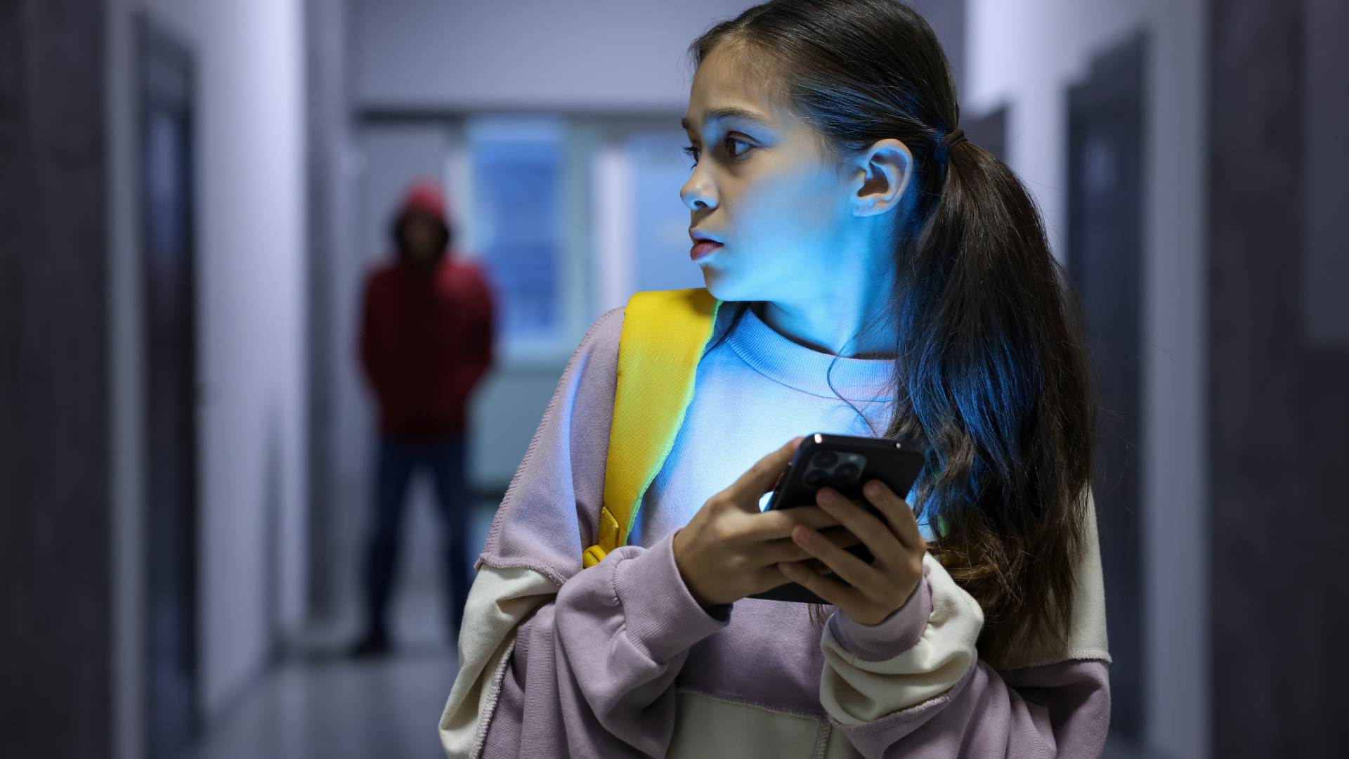 A young person looking back with a worried expression while holding a glowing phone, with a figure following in a hallway.