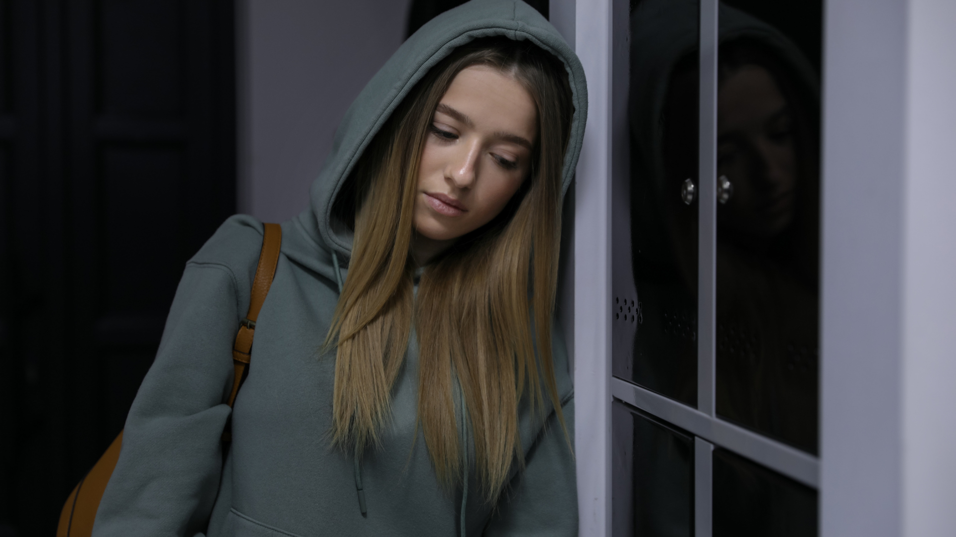 A person in a sage green hoodie leans against a white doorframe, looking down with a somber expression.