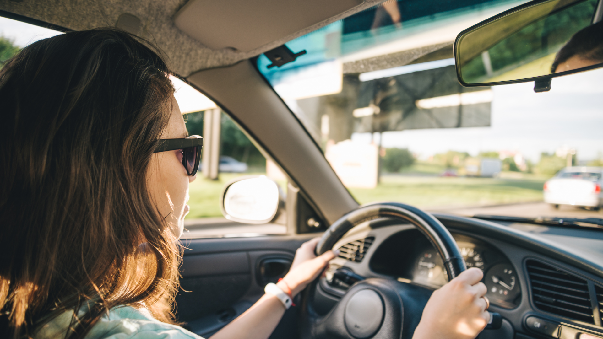 A person wearing sunglasses grips the steering wheel of a car while driving on a road during the day.