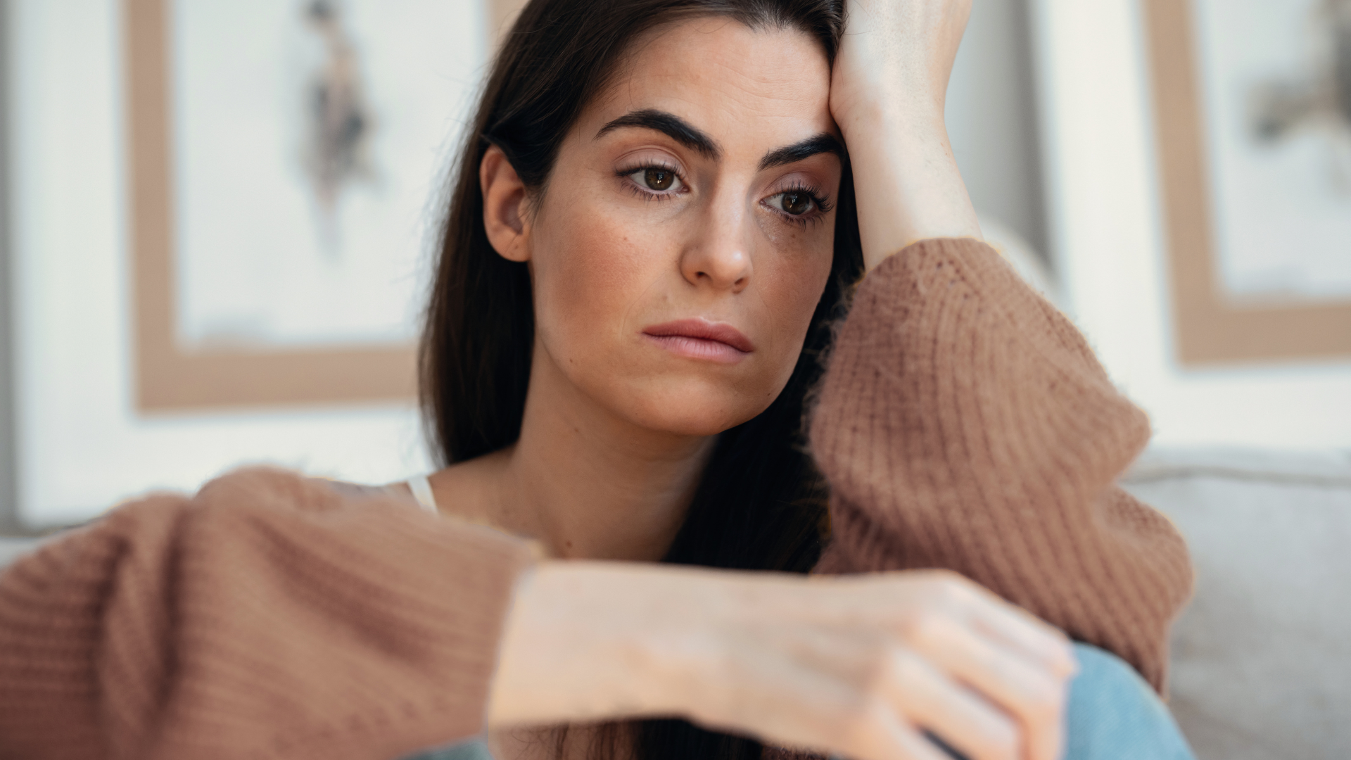 A person with a thoughtful expression rests their head on their hand while wearing a brown sweater in a soft indoor setting.