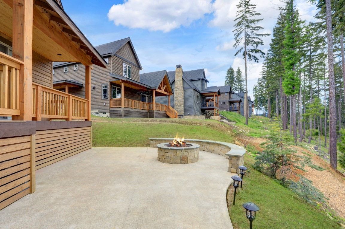 Wooden cabin patio with stone fire pit, overlooking a grassy slope and tall pine trees