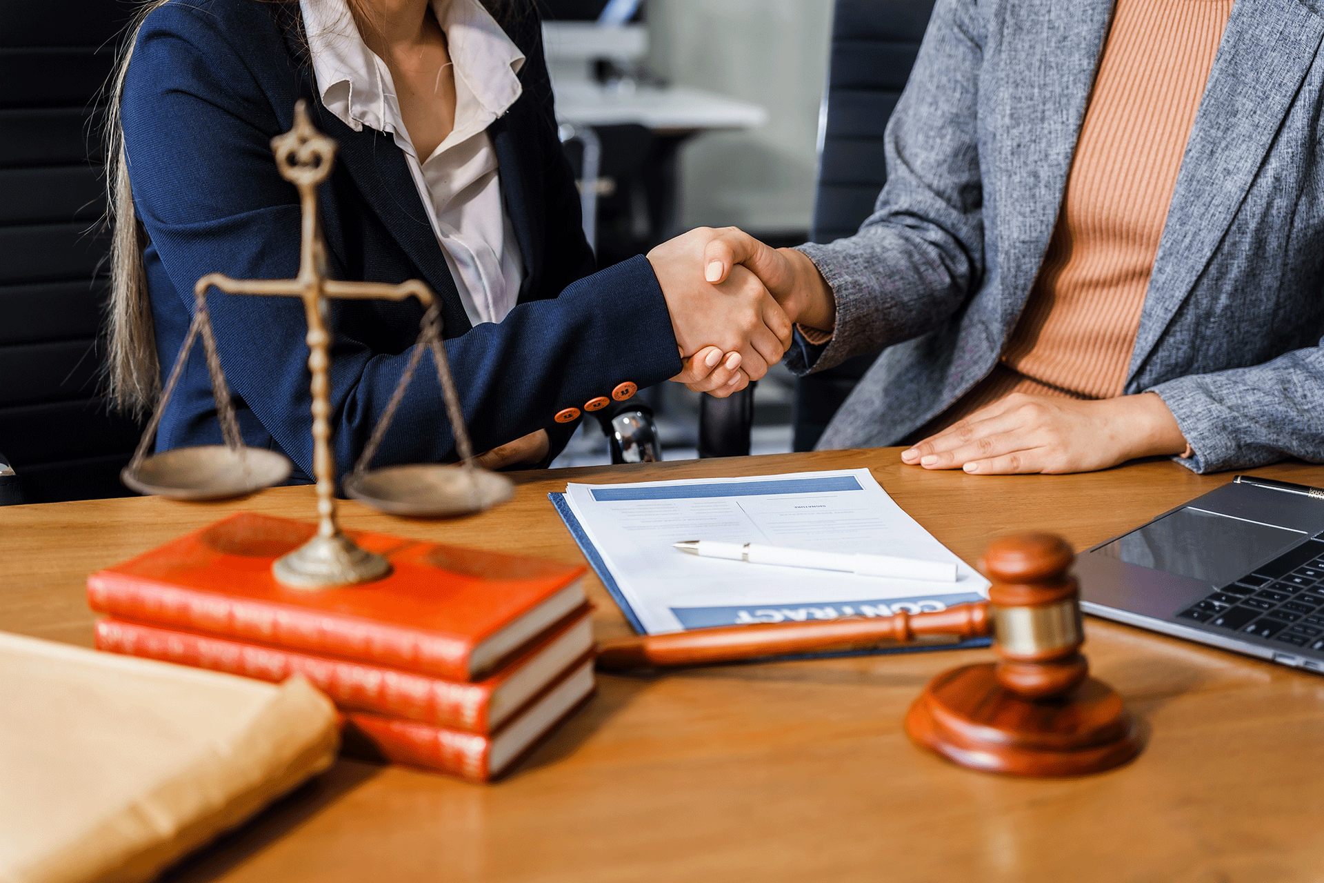 Two people shaking hands over a contract on a desk, next to law books, scales, and gavel.