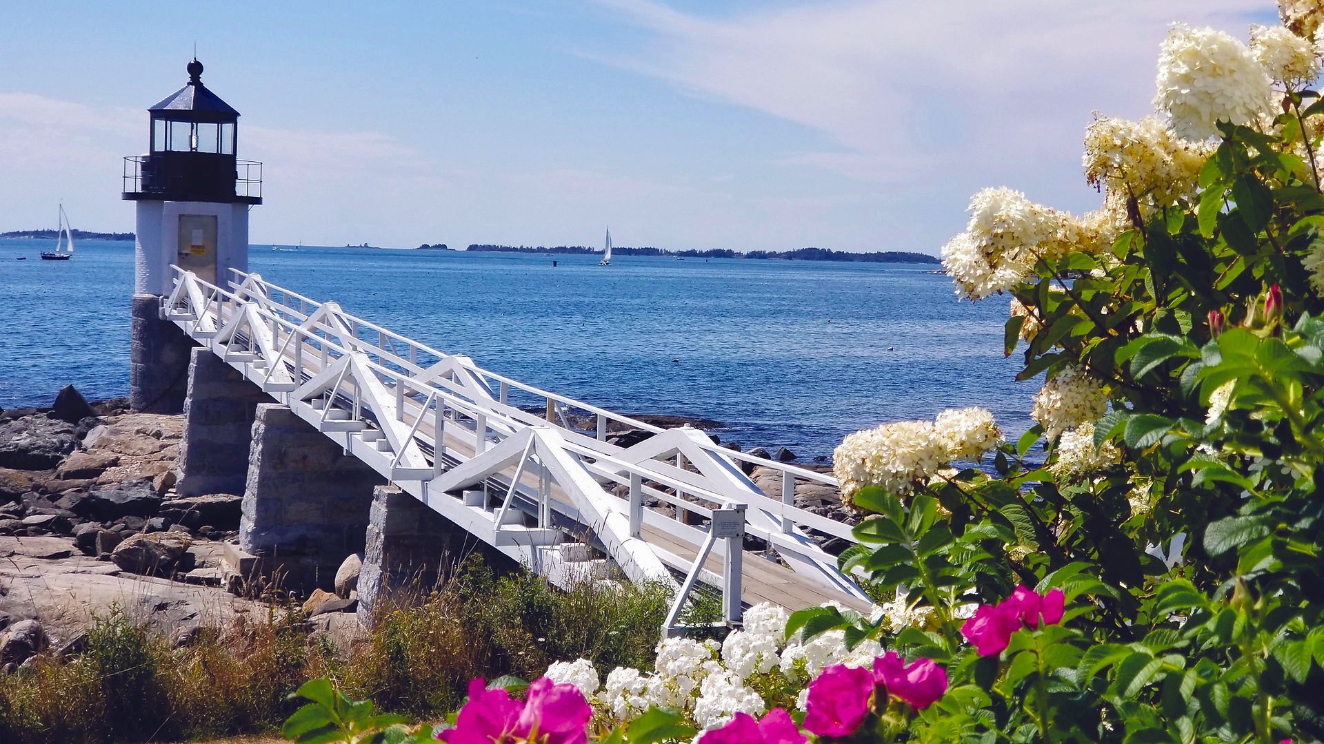 Lighthouse on a stone pier, connected by a white wooden bridge, blue ocean, and flowers in bloom.