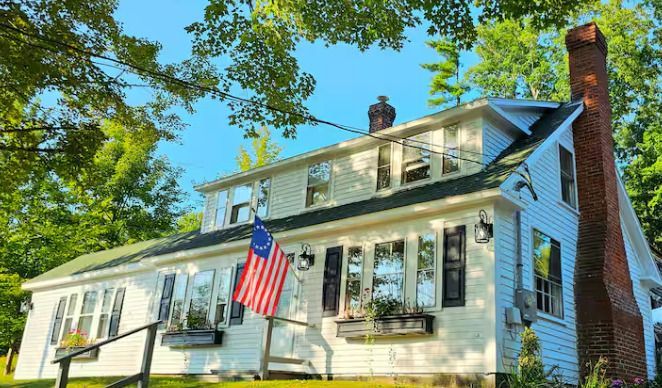 White colonial house with an American flag, brick chimney, and trees under a bright blue sky