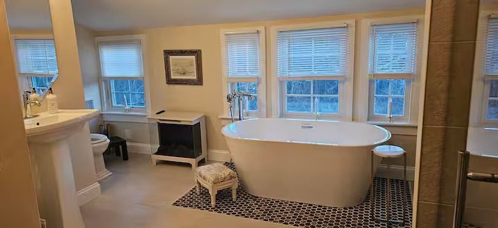 Bright bathroom with a white bathtub, patterned black-and-white tile, and windows with blinds