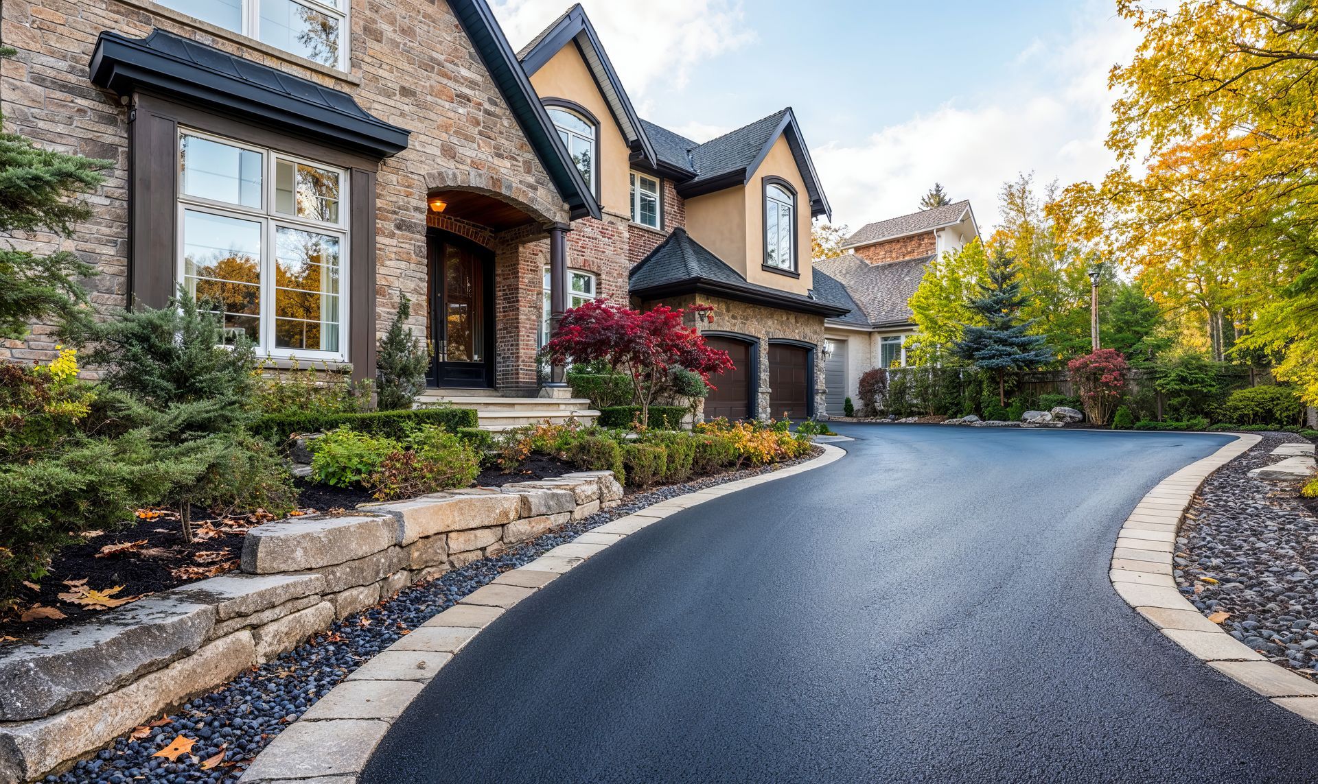 A modern stone house with a curved asphalt driveway, bordered by a stone retaining wall and lush landscaping.