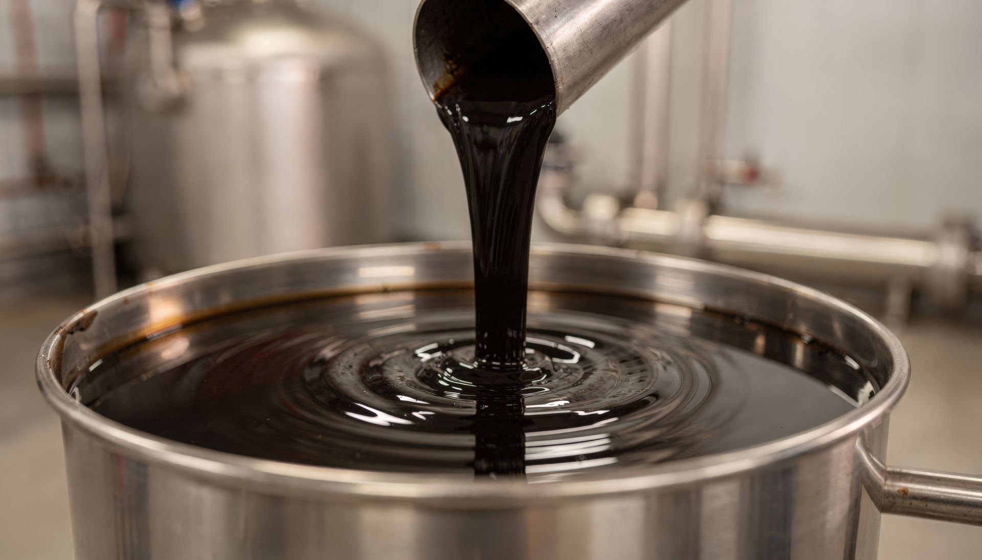 Dark oil pouring from a metal spout into a stainless steel container in an industrial setting